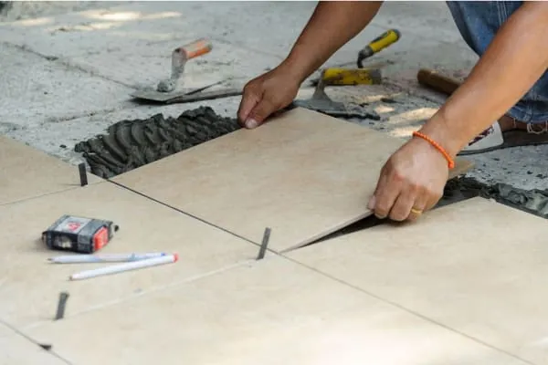 Person laying tan tiles on a concrete surface, using spacers and a trowel outdoors.