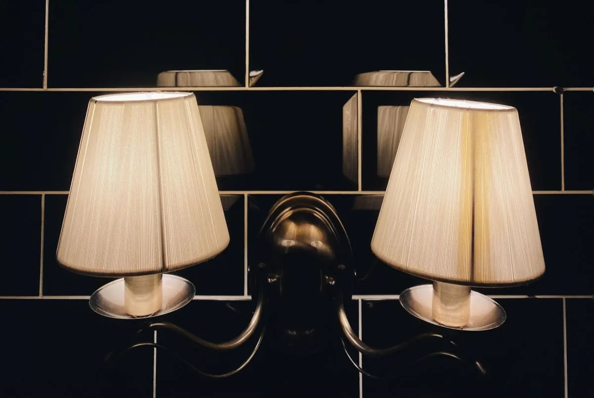 Pair of cream pleated wall sconces on a dark tiled wall, with a central brass fixture between them.