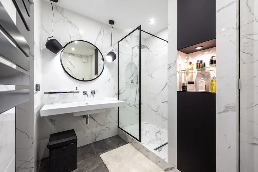 Modern bathroom with marble walls, round mirror, white vanity, glass shower, and black trash bin