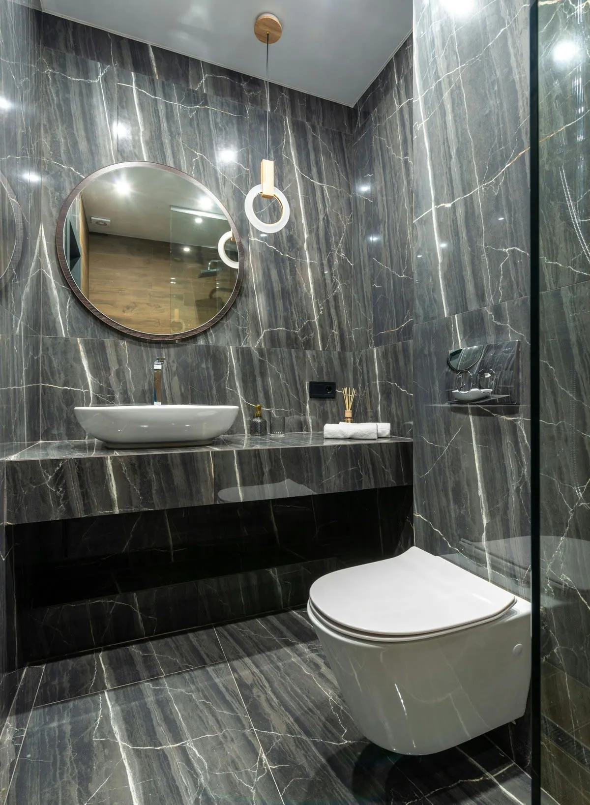 A modern bathroom with dark marble walls and floors. It features a round mirror, floating sink, and a white toilet.