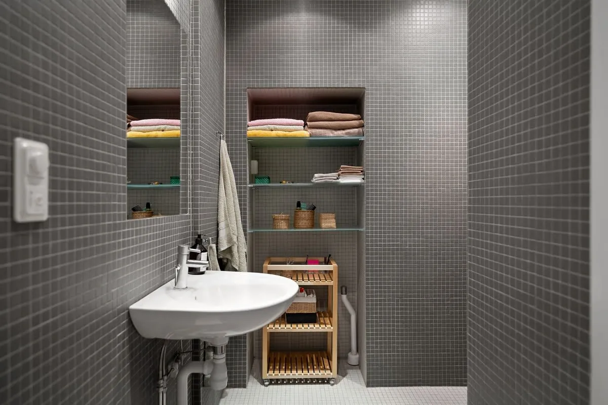 Small tiled bathroom with a white sink, open storage shelves, folded towels, and a wicker hamper.