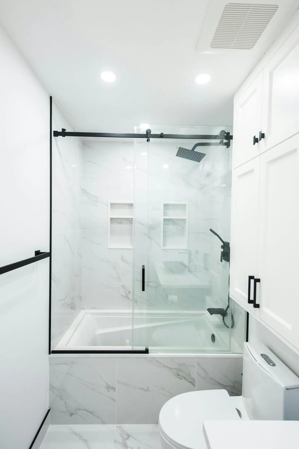 Modern white bathroom with marble tile, black shower door, fixtures, and tall white cabinets.