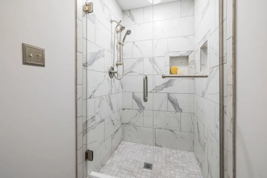 Marble-tiled walk-in shower with glass door, chrome fixtures, and built-in shelf with soap