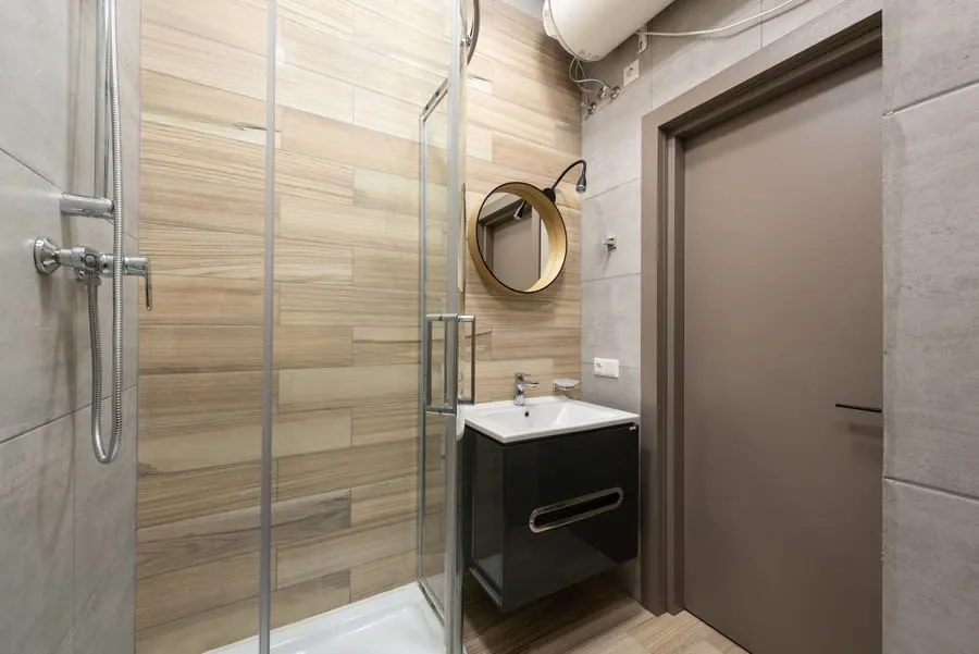 Modern bathroom with glass shower, wood-look tile, black vanity, round mirror, and closed door