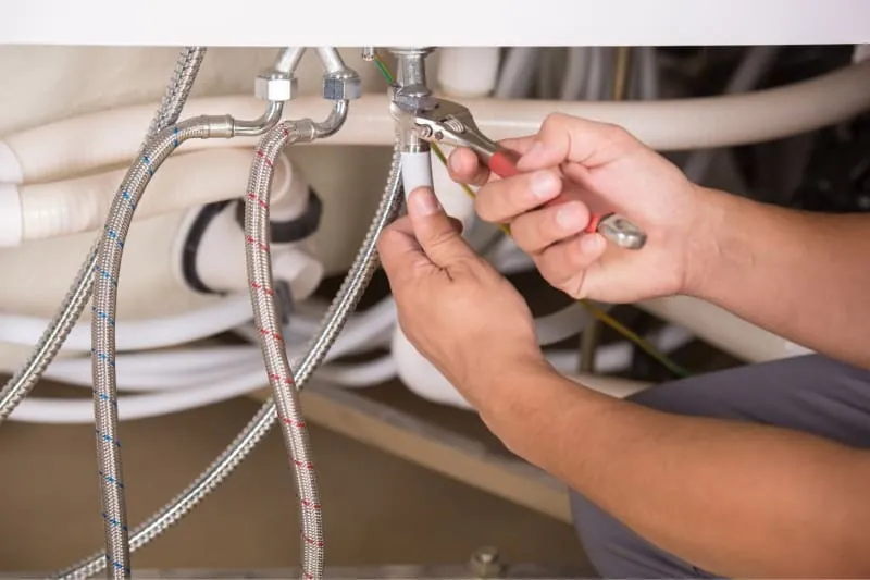 A plumber's hands using pliers on plumbing pipes beneath a sink.