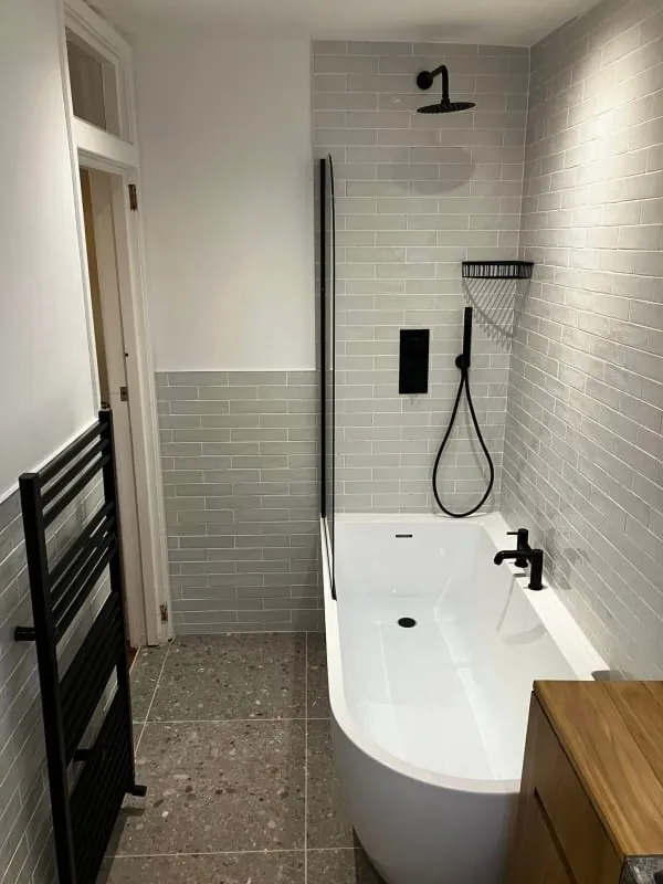 Small bathroom with gray brick-like tiles, black fixtures, white tub, and a wooden cabinet.