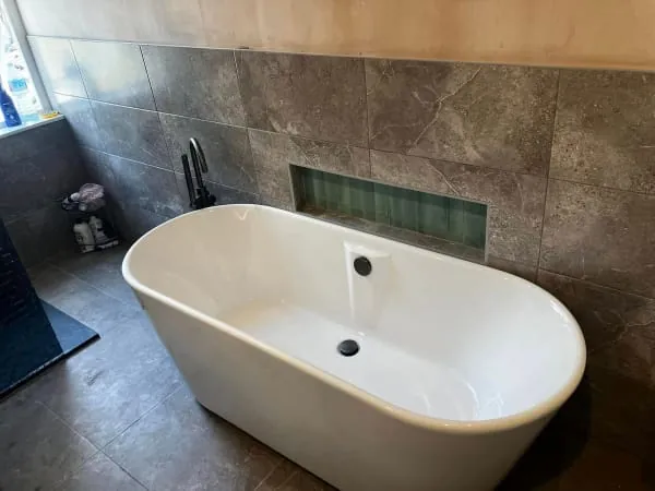 Freestanding white bathtub against a gray tiled wall with a built-in shelf and dark faucet.