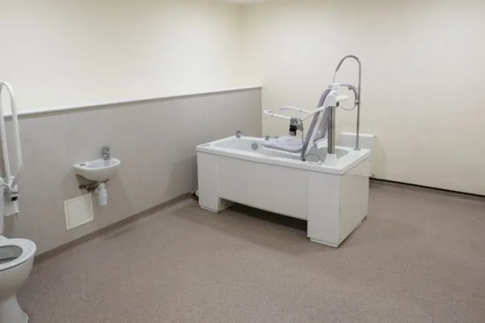 Accessible bathroom with bathtub-style chair lift, wall-mounted sink, and toilet; beige and white colors.