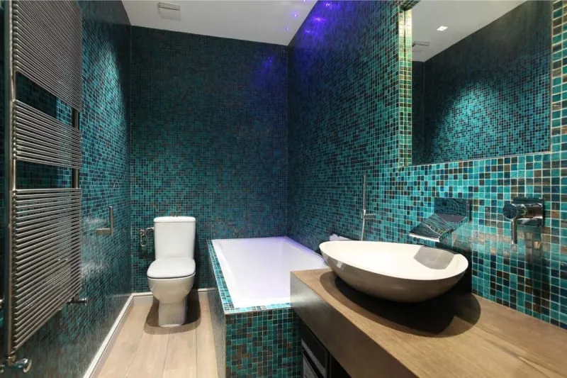 Bathroom with teal mosaic tile walls, white toilet, tub, and sink, and a wooden vanity.