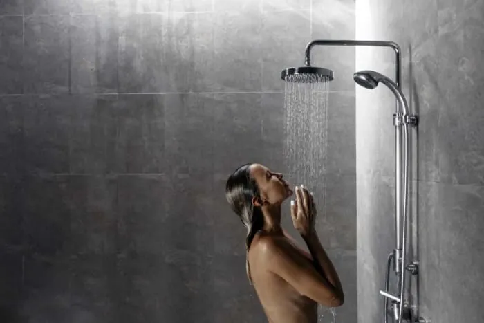 Woman in shower, head tilted back, arms raised to face, water streaming down. Gray tiled walls.