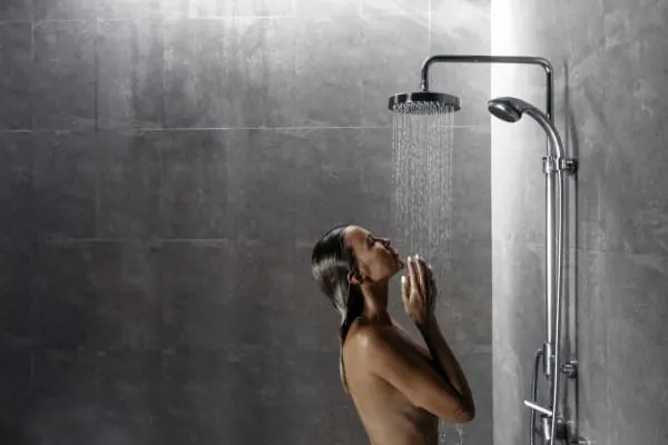 Woman in a shower with water cascading onto her face, hands clasped. Gray tiled walls, silver fixtures.