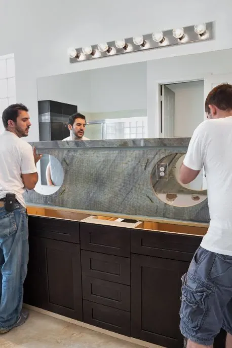 Two men install a bathroom vanity countertop. The dark brown cabinet is beneath a gray countertop with two sink cutouts.