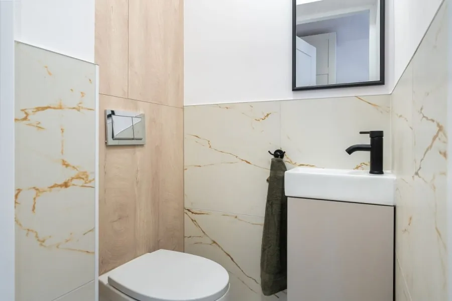 Small modern bathroom with marble walls, white sink, black faucet, and a toilet beside a wooden panel.